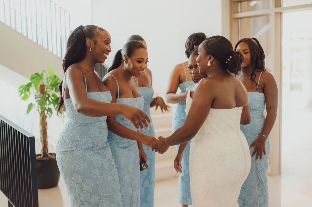 Bride in white gown holding hands with bridesmaids in light blue lace dresses during wedding preparation.
