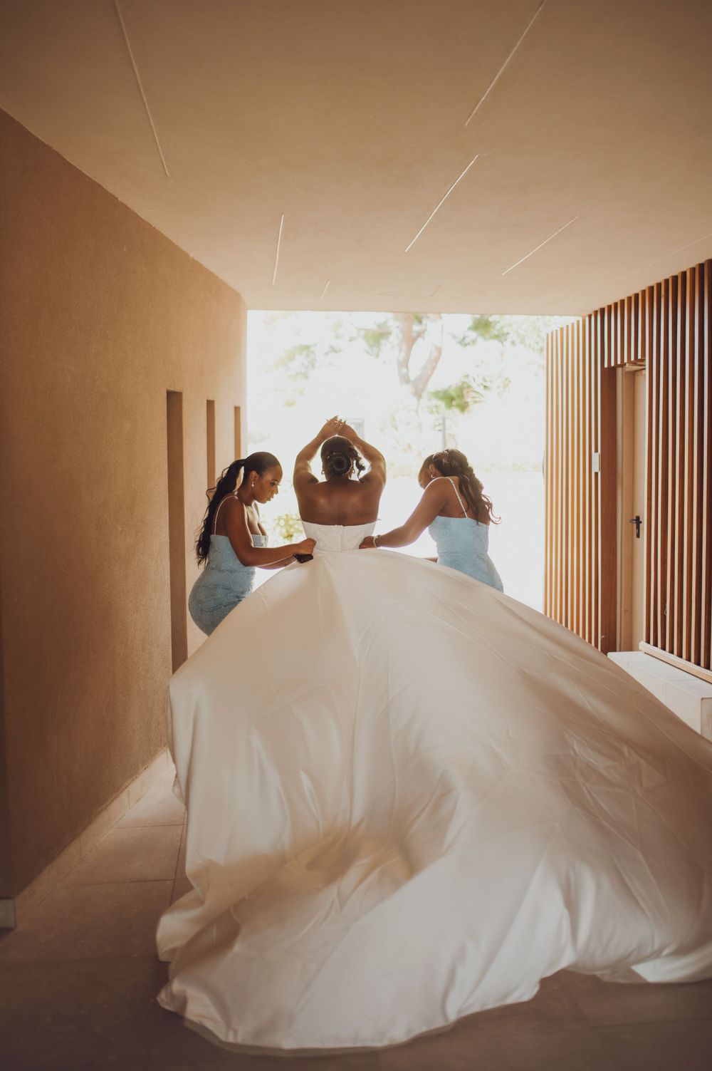 Bride in flowing white gown assisted by two bridesmaids in blue dresses in a modern hallway.