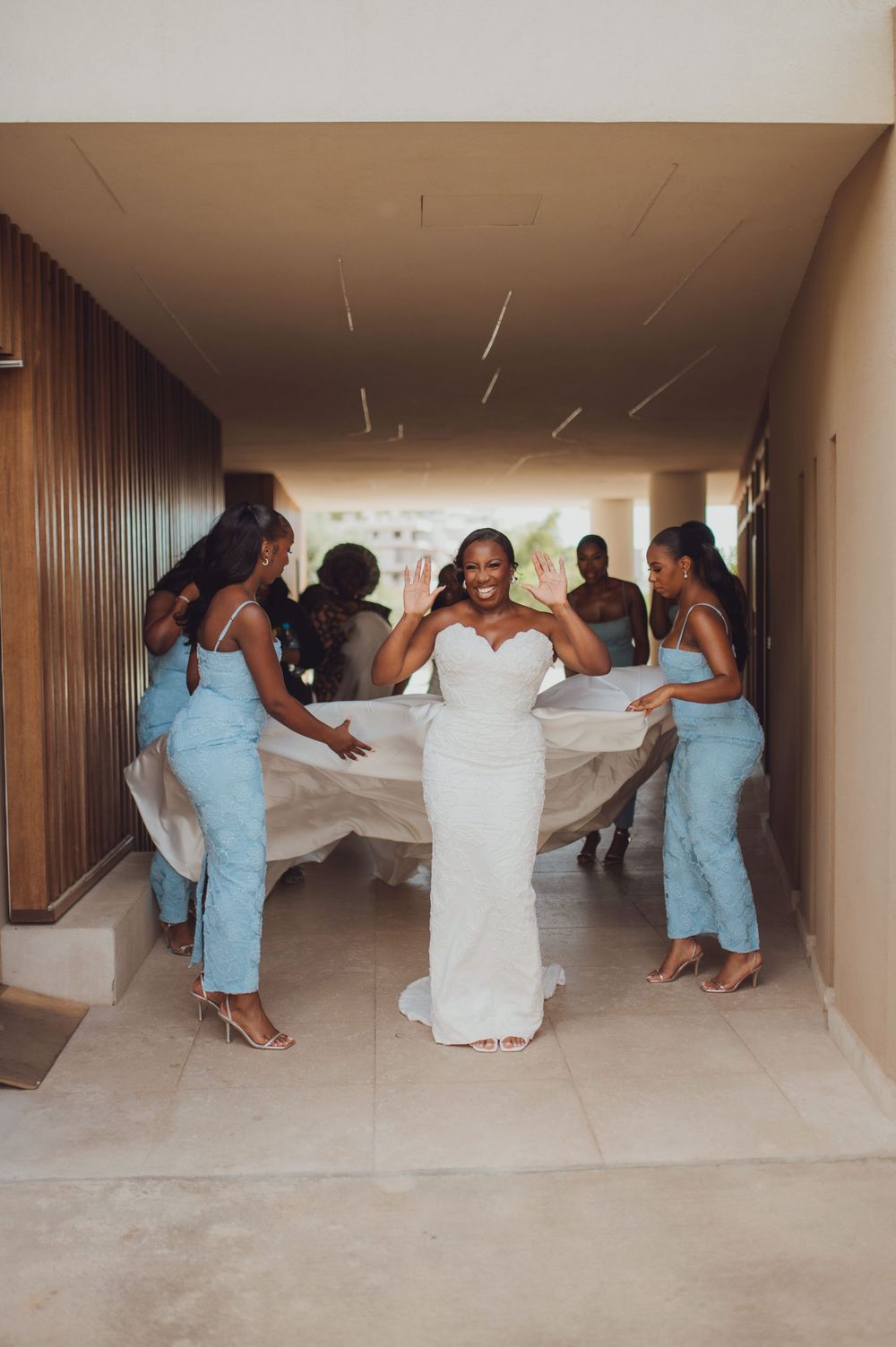 Bride in white gown smiles as bridesmaids in blue dresses hold her train in elegant hallway.