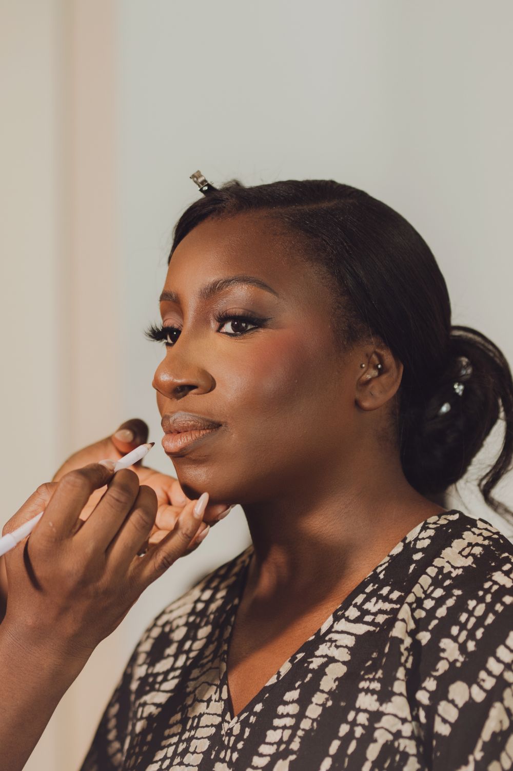 A Black woman applies lipstick while wearing a patterned robe, showcasing elegant makeup with long lashes and rosy cheeks.