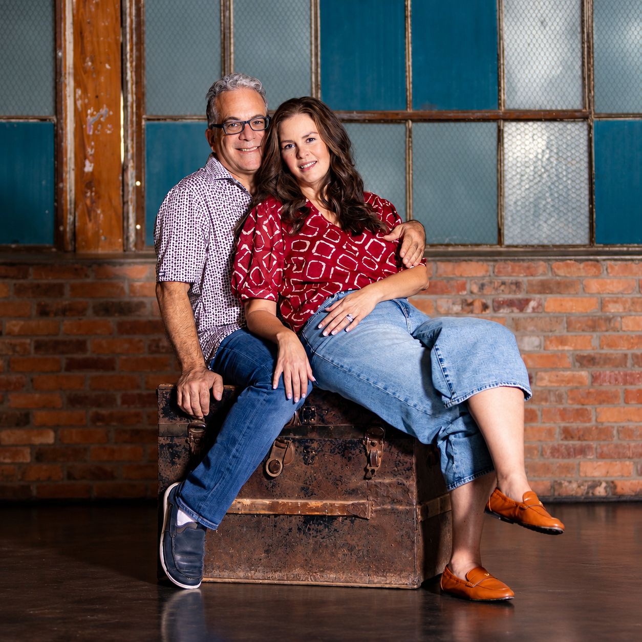 A couple smiles warmly while posing together on a vintage trunk against a brick wall backdrop.