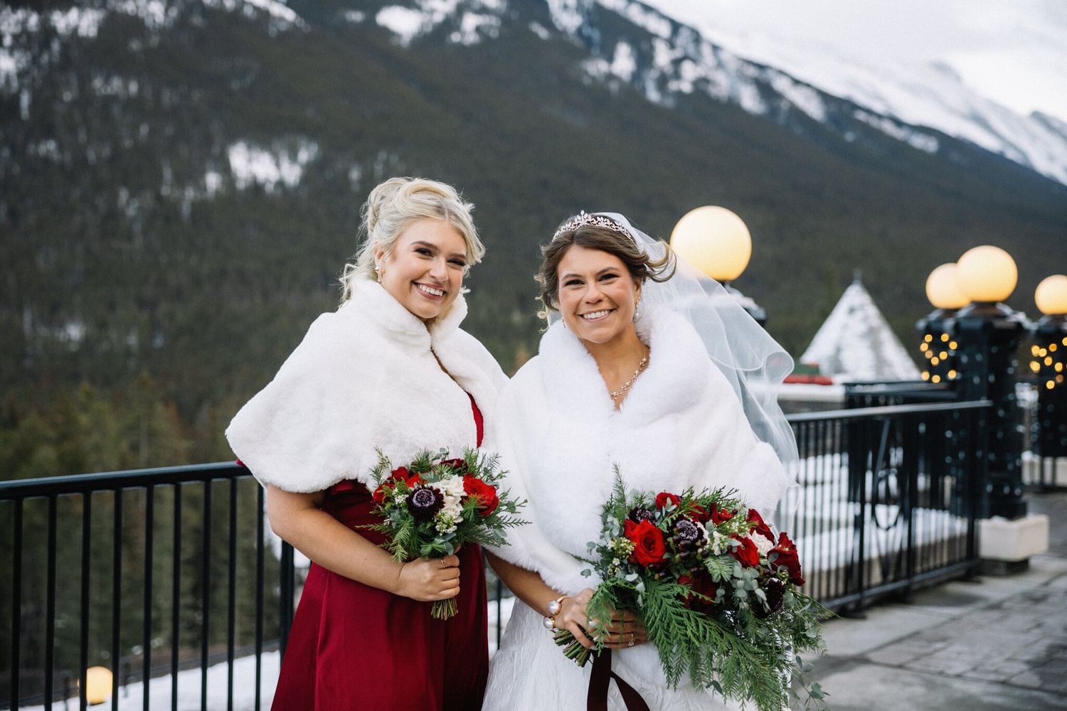 Bride in a white wrap and bridesmaid in a red dress smile with red rose bouquets on a snowy Banff terrace