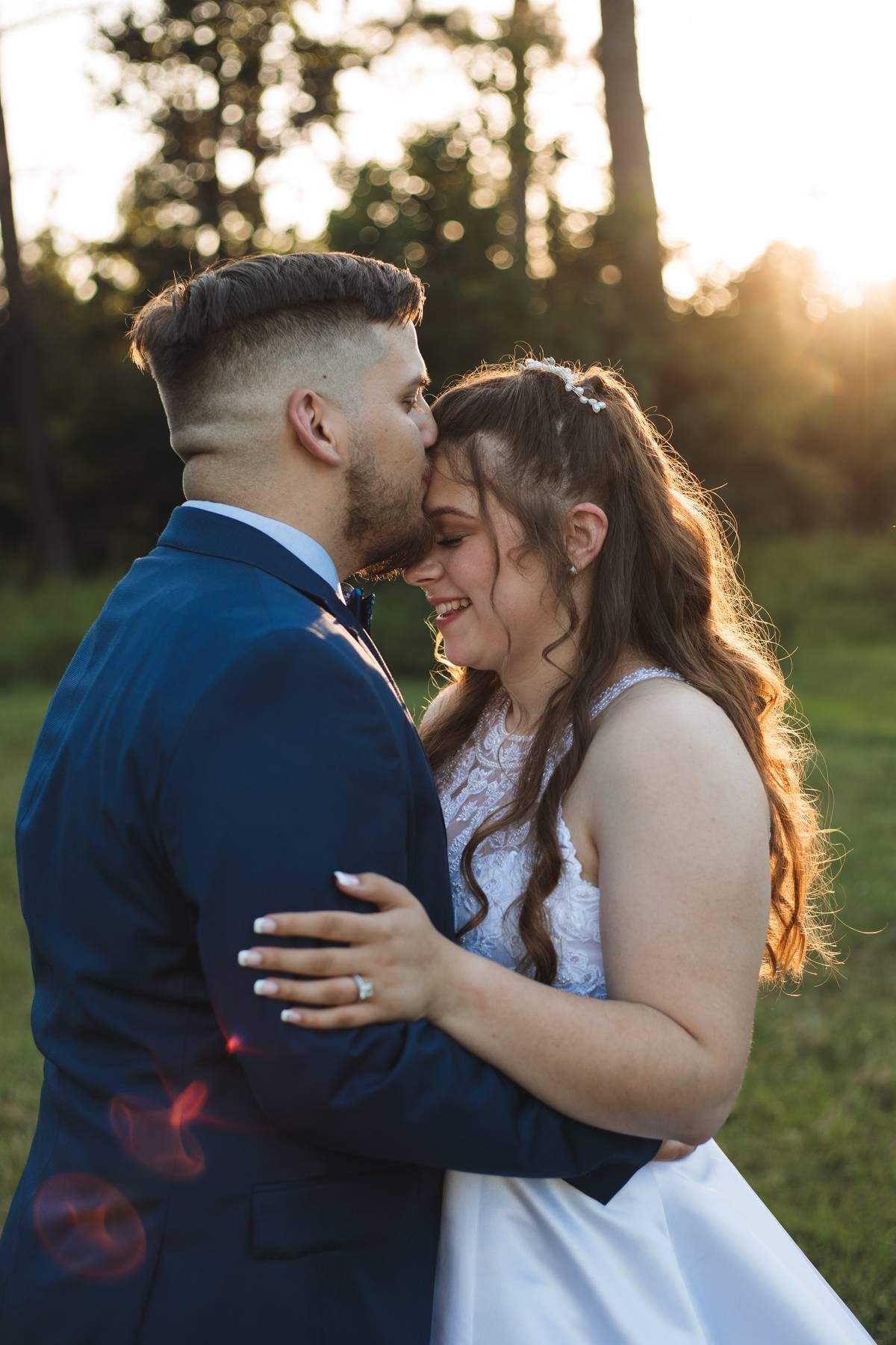 Groom in a navy suit kisses the forehead of his smiling bride in a white dress at golden hour in a sunlit field.