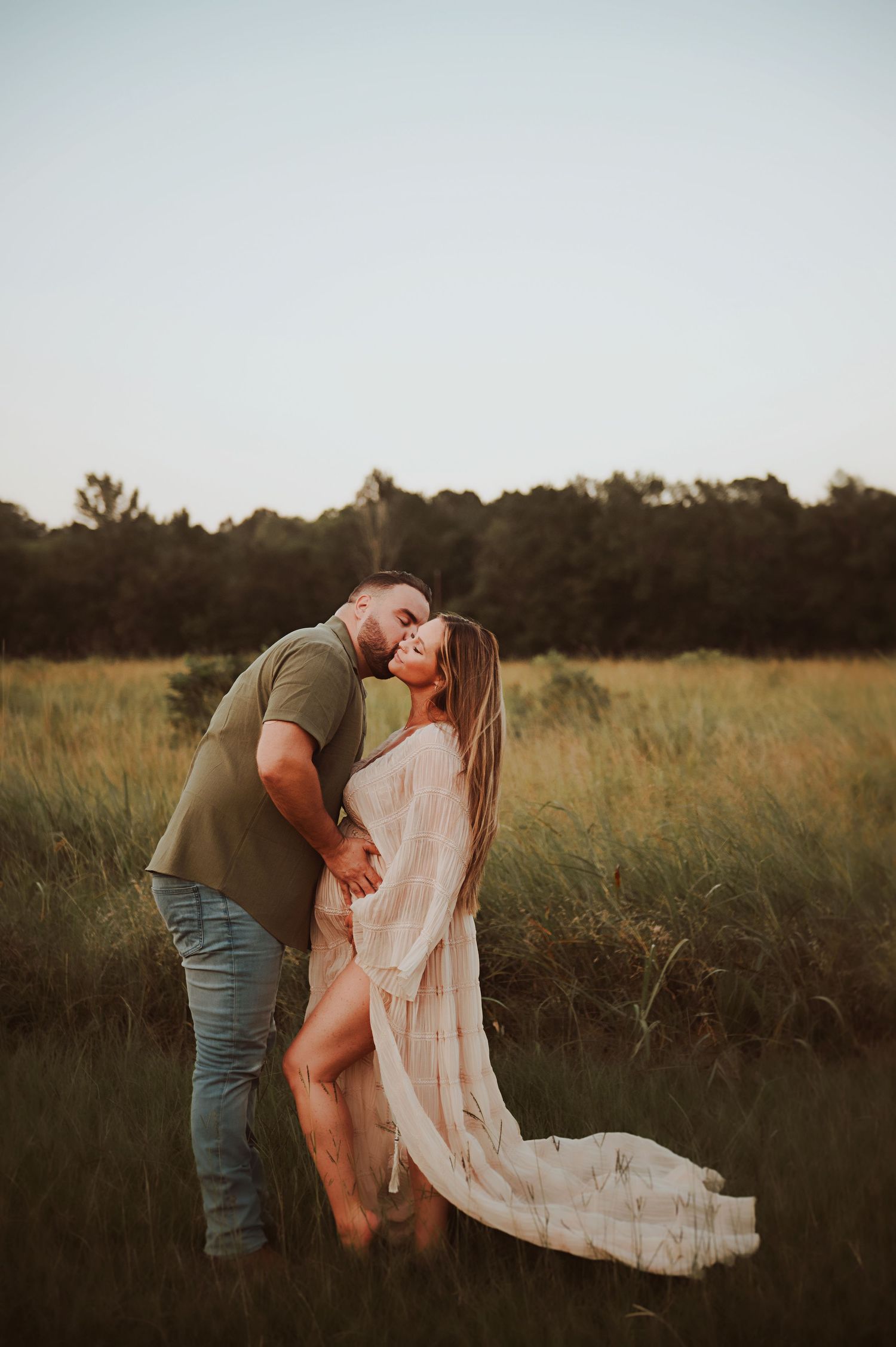 Couple sharing a tender kiss in a golden grassy field at sunset, she wears a flowing cream gown, he wears an olive tee and jeans.