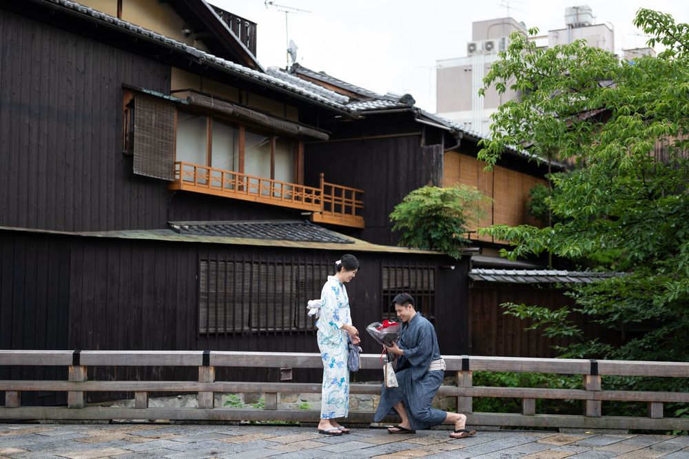Couple in traditional Japanese kimono and yukata pose beside a wooden railing in front of a historic dark-timber machiya house in Kyoto.