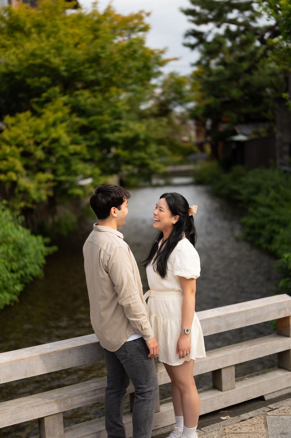 A smiling couple stands face to face on a wooden bridge over a calm stream, surrounded by lush green trees.