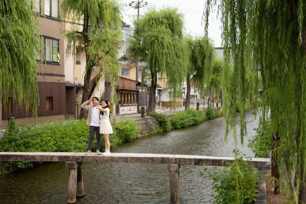 Couple poses for a photo on a stone bridge over a calm canal lined with weeping willow trees and traditional buildings in Kyoto.