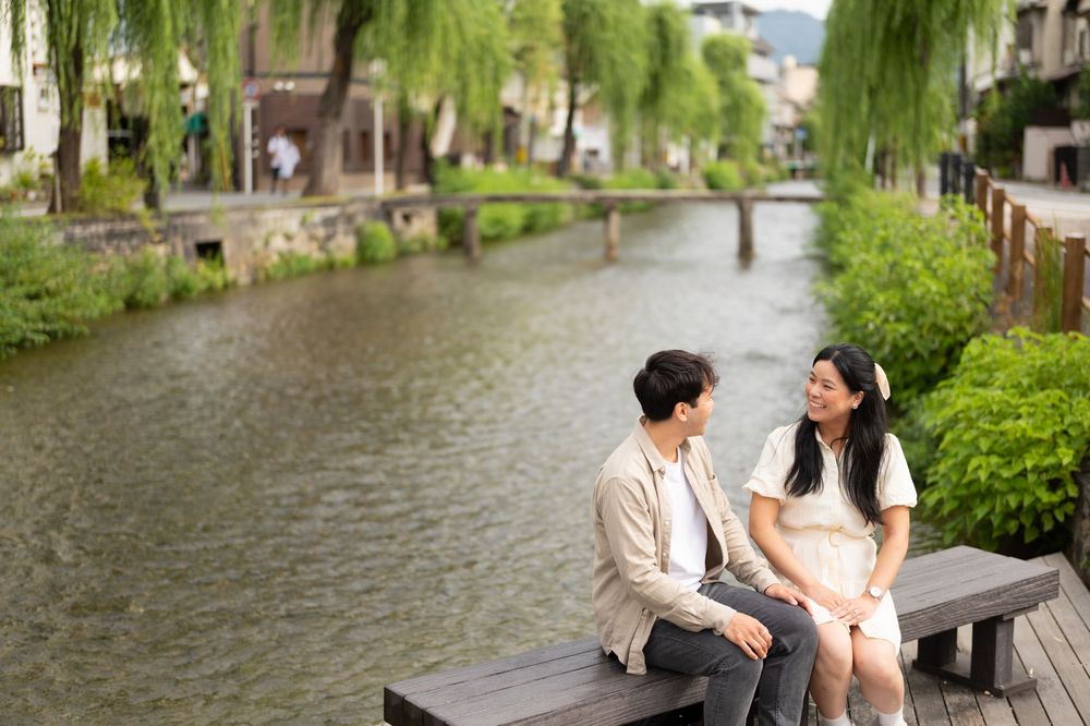 Smiling couple sits on a wooden bench beside a calm canal lined with willow trees and lush greenery in a charming historic district.