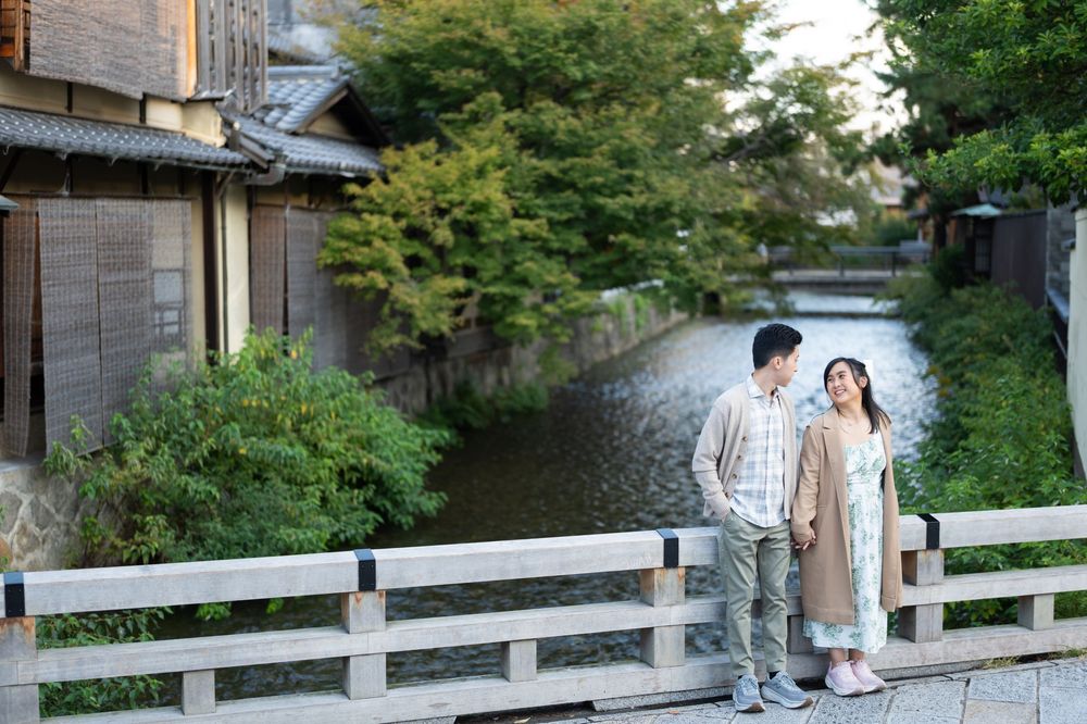 Couple standing together on a wooden bridge over a calm canal, surrounded by lush green trees and traditional Japanese buildings.