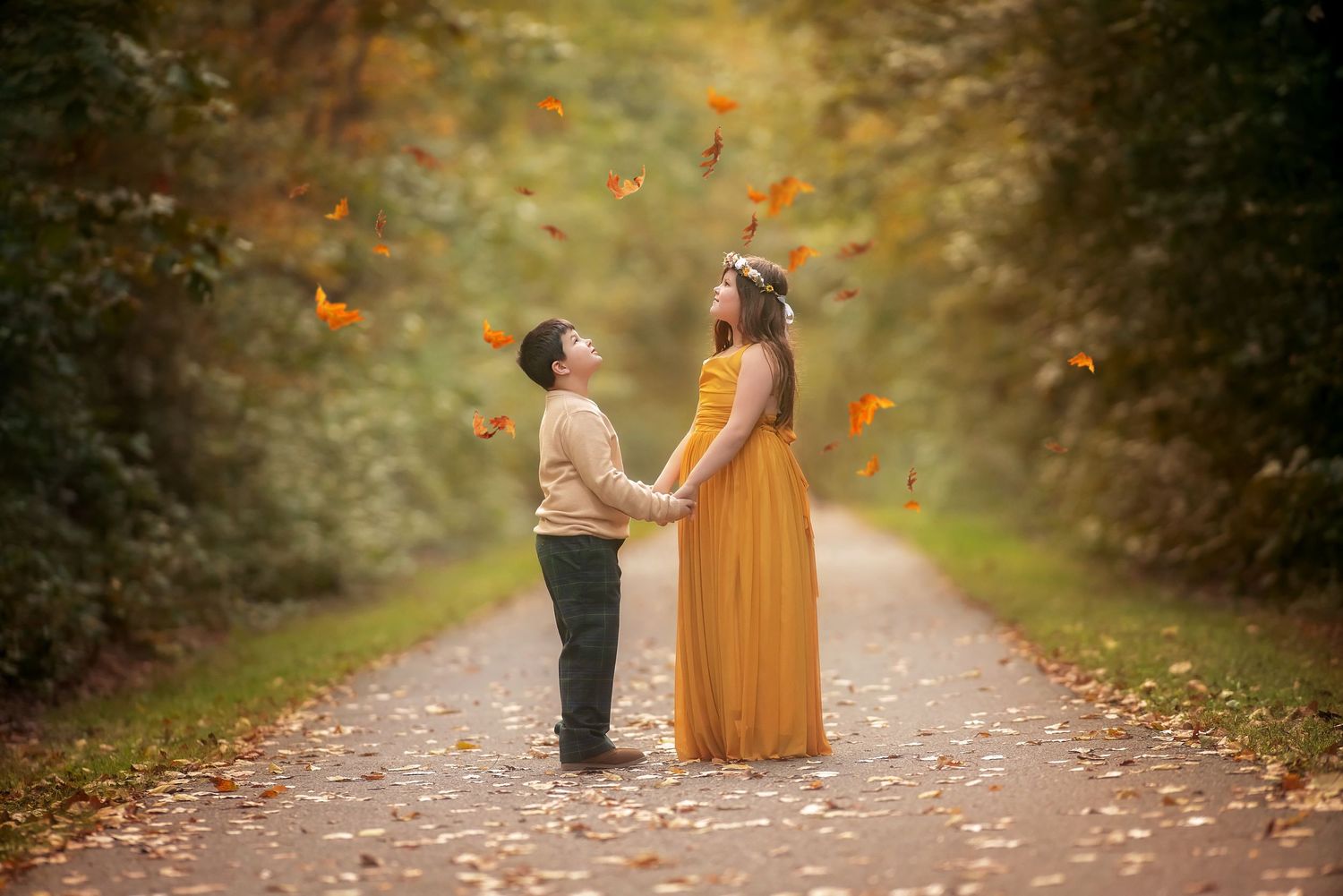Two children holding hands on a leaf-covered forest path in autumn, one in a long yellow dress with a floral crown, as leaves fall.