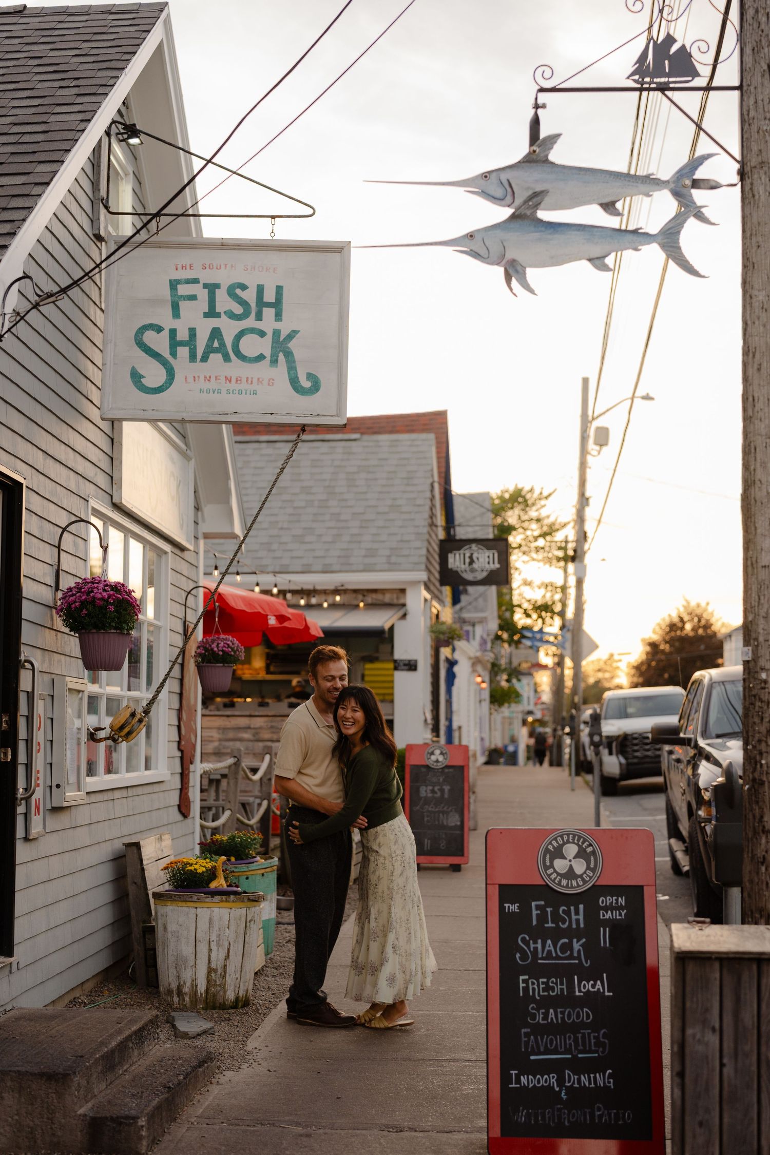 Couple embracing outside Fish Shack seafood restaurant at golden hour, with decorative swordfish hanging above.