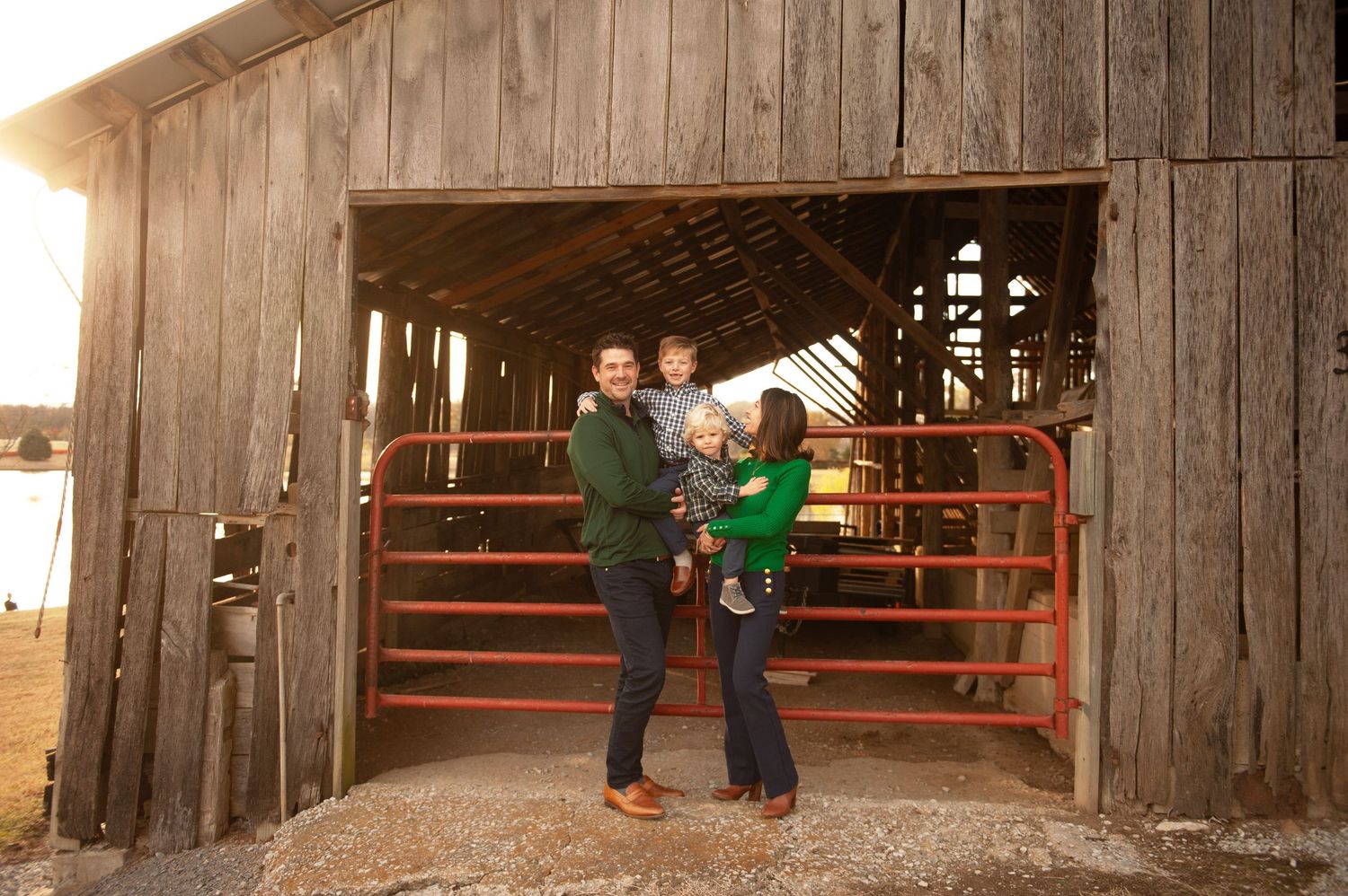 Family of three posing in front of a rustic wooden barn with a red gate at golden hour.