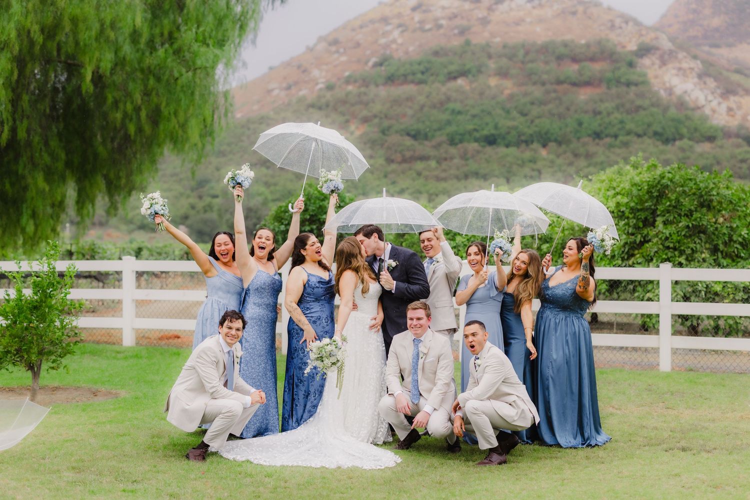Bride and groom kiss surrounded by bridesmaids in blue dresses and groomsmen holding white umbrellas on a grassy lawn.
