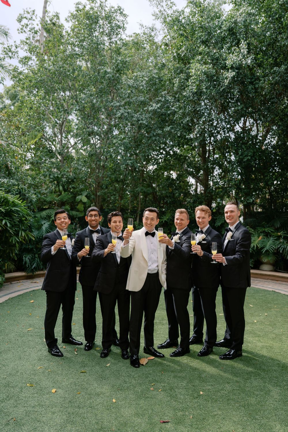 Groom in white tuxedo jacket raises champagne toast with groomsmen in black suits outdoors.