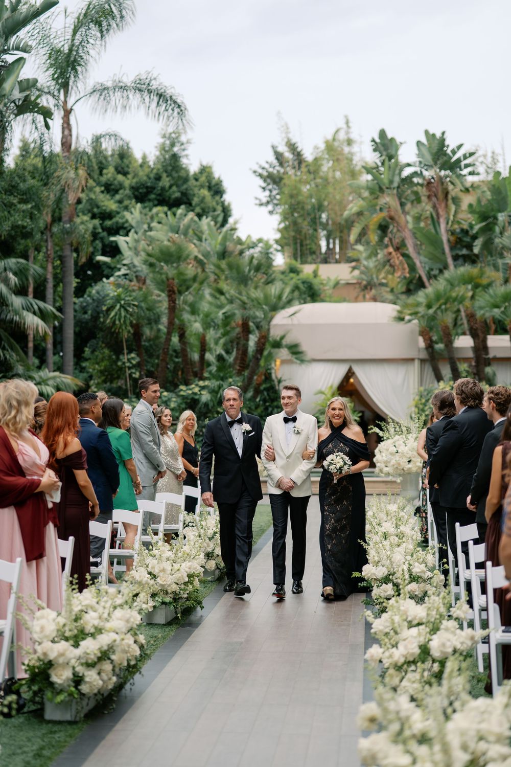 Groom walks down floral-lined aisle with parents at elegant outdoor wedding ceremony surrounded by lush tropical greenery.