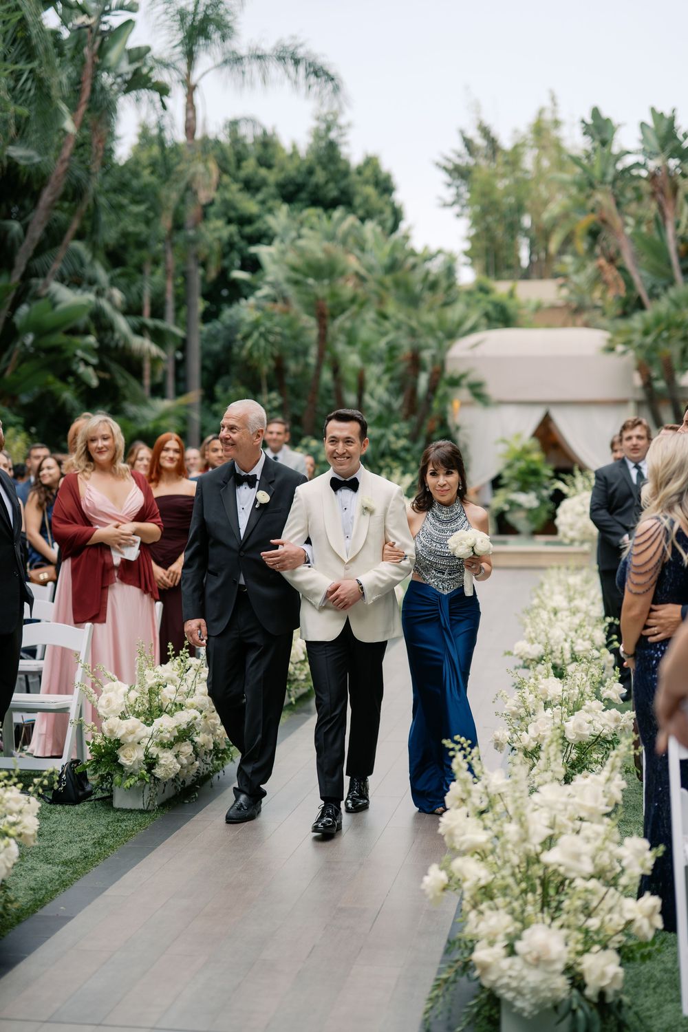 Groom walks down aisle with parents at outdoor wedding ceremony, flanked by white floral arrangements and lush tropical greenery.