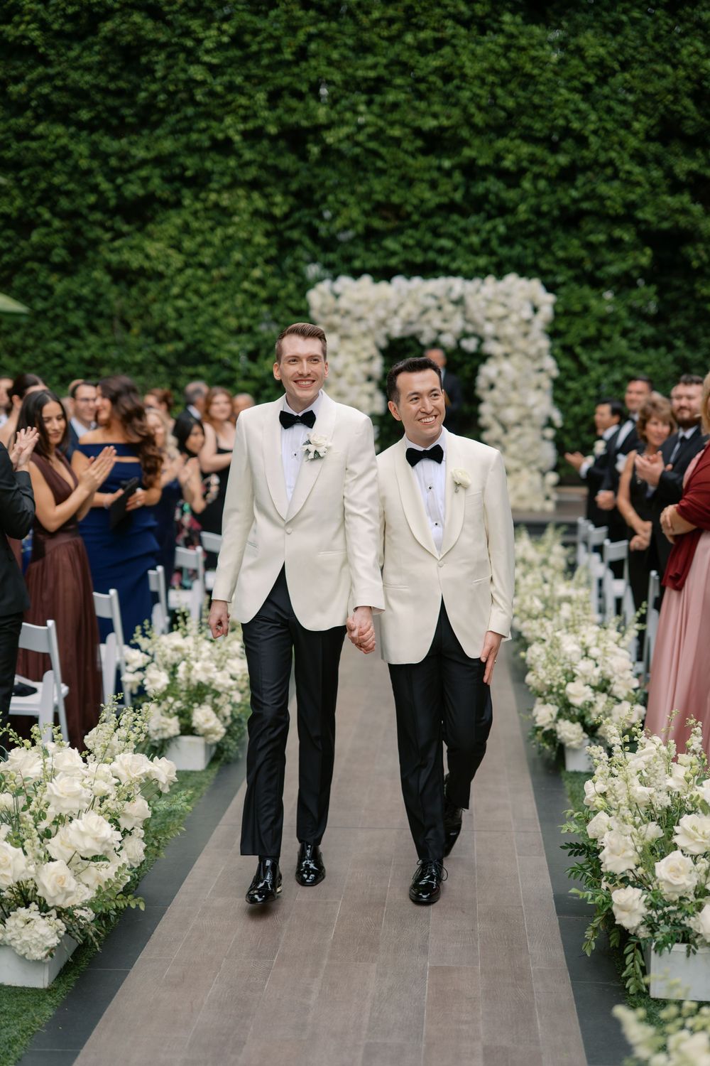 Two grooms in white tuxedo jackets walk down floral-lined aisle at elegant outdoor wedding ceremony.