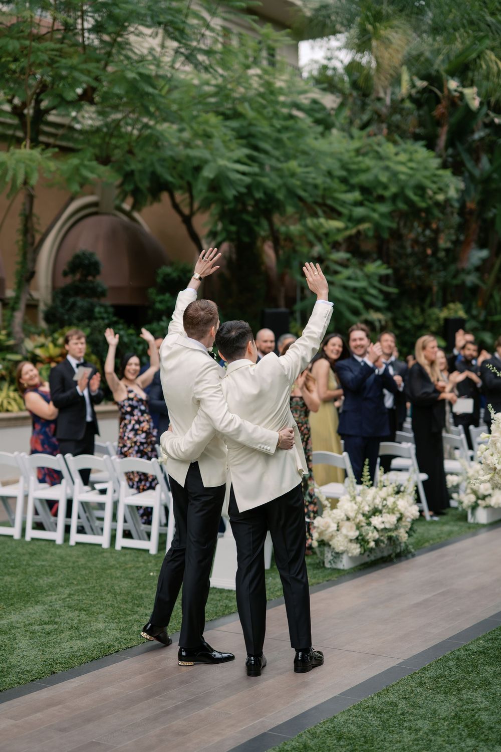 Two grooms walk down the aisle waving to guests at an outdoor wedding ceremony surrounded by lush greenery.