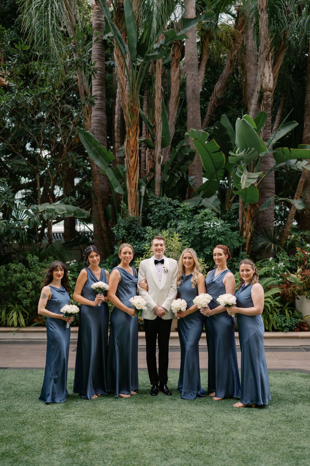 Groom in white tuxedo jacket poses with six bridesmaids in dusty blue gowns holding white bouquets outdoors.