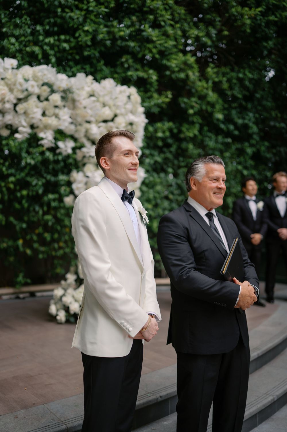 Groom in white tuxedo jacket stands with officiant before floral arch at elegant outdoor wedding ceremony.