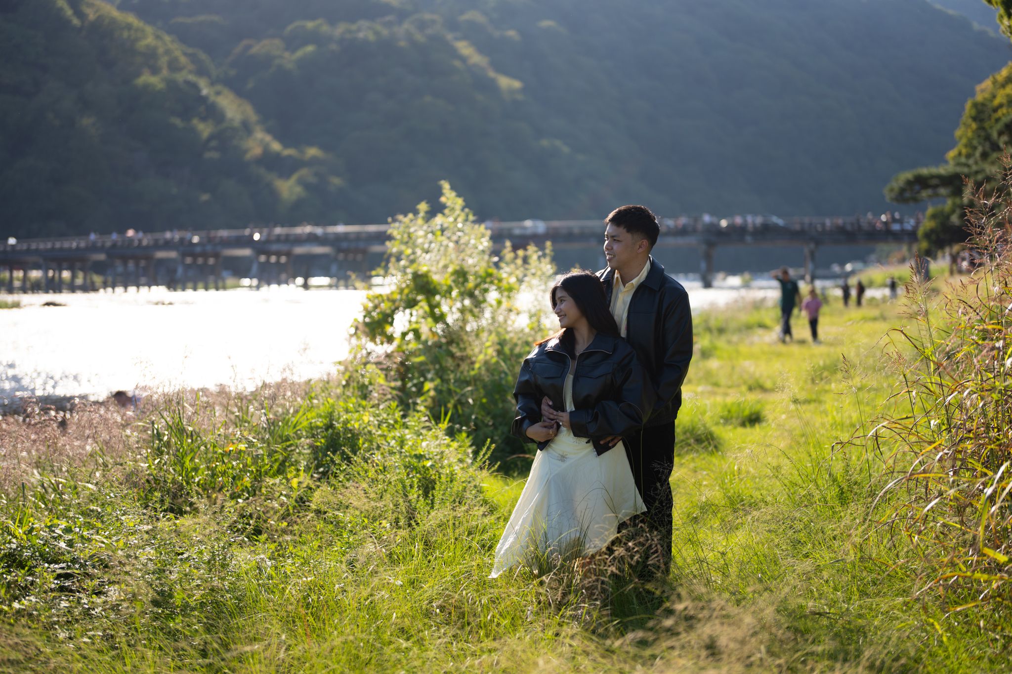 Couple in dark clothing and a white dress embracing in a grassy riverside field with a long bridge and forested mountains in the background.