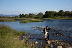 Couple in formal attire stands on rocky riverbed in shallow water, surrounded by green grassy banks, trees and a blue sky.