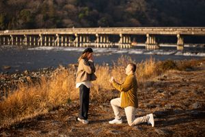 Man kneeling on one knee proposing to a woman covering her mouth in surprise on a golden grassy riverbank with a long bridge behind them.