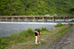 Couple embracing on a grassy riverbank beside a wide shallow river, with a long bridge and lush green forested hills in the background.