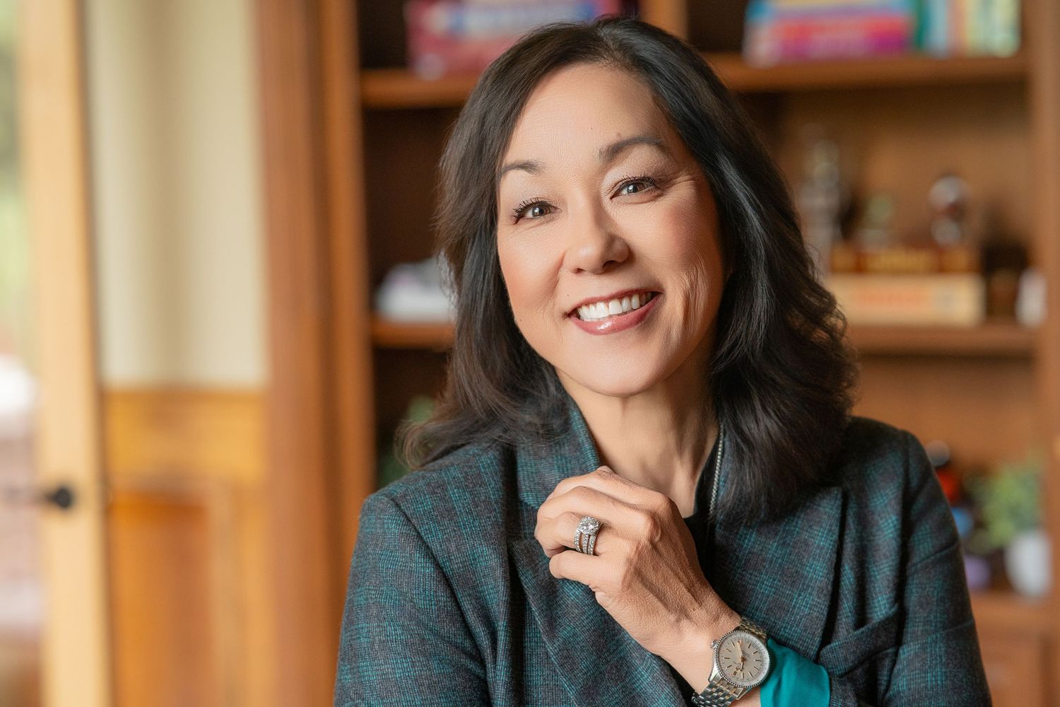 Smiling Asian woman with dark hair in teal blazer poses indoors near wooden shelves in warm, professional setting.