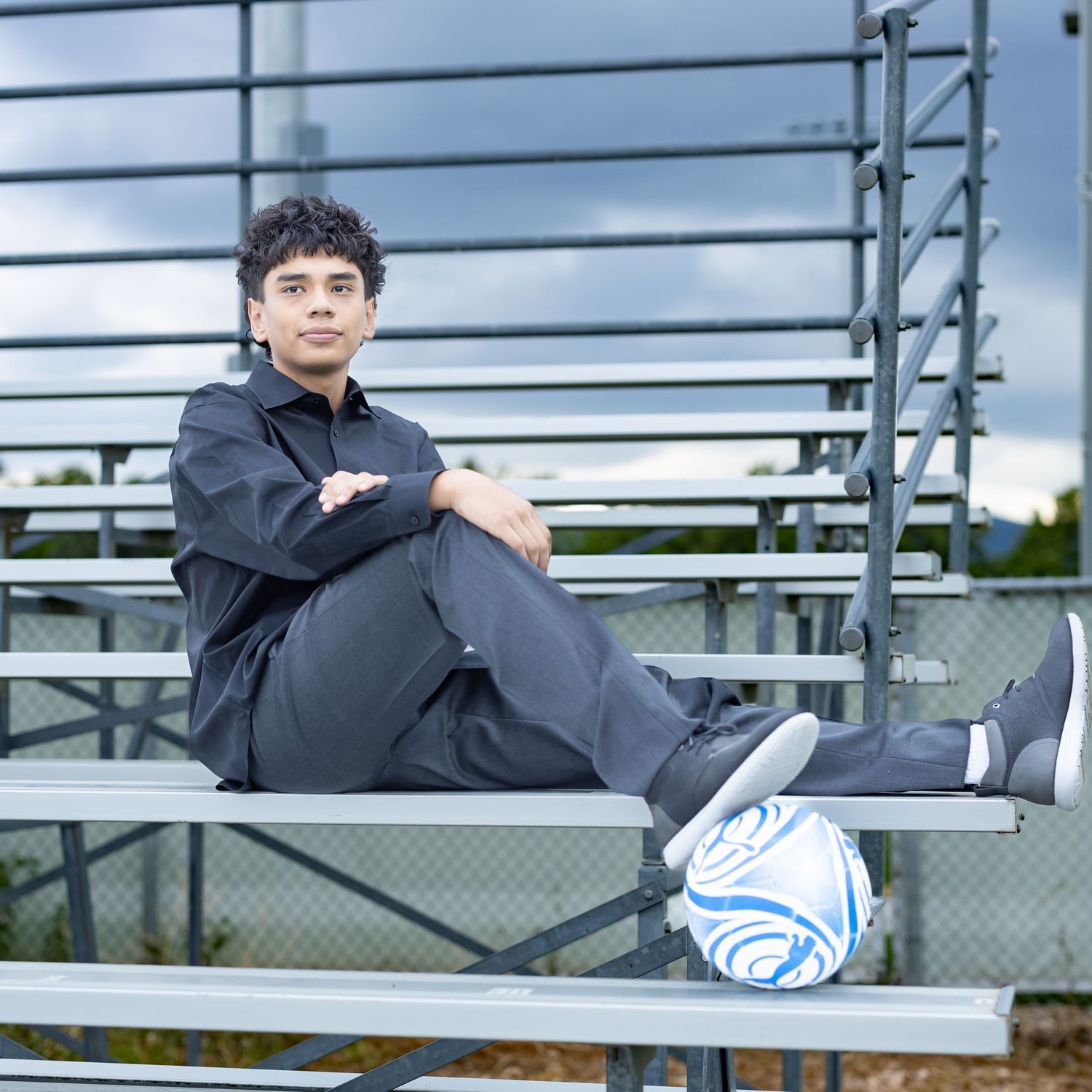 Teen boy in dark outfit sits on bleachers with soccer ball, posing for senior portrait outdoors.
