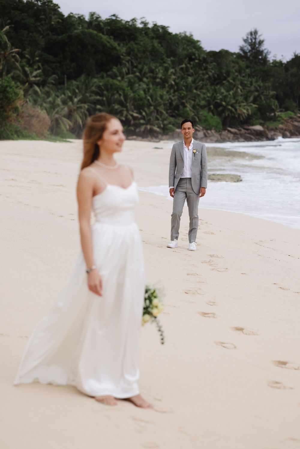 Bride in white strapless gown stands on sandy beach as groom approaches from behind in gray suit.