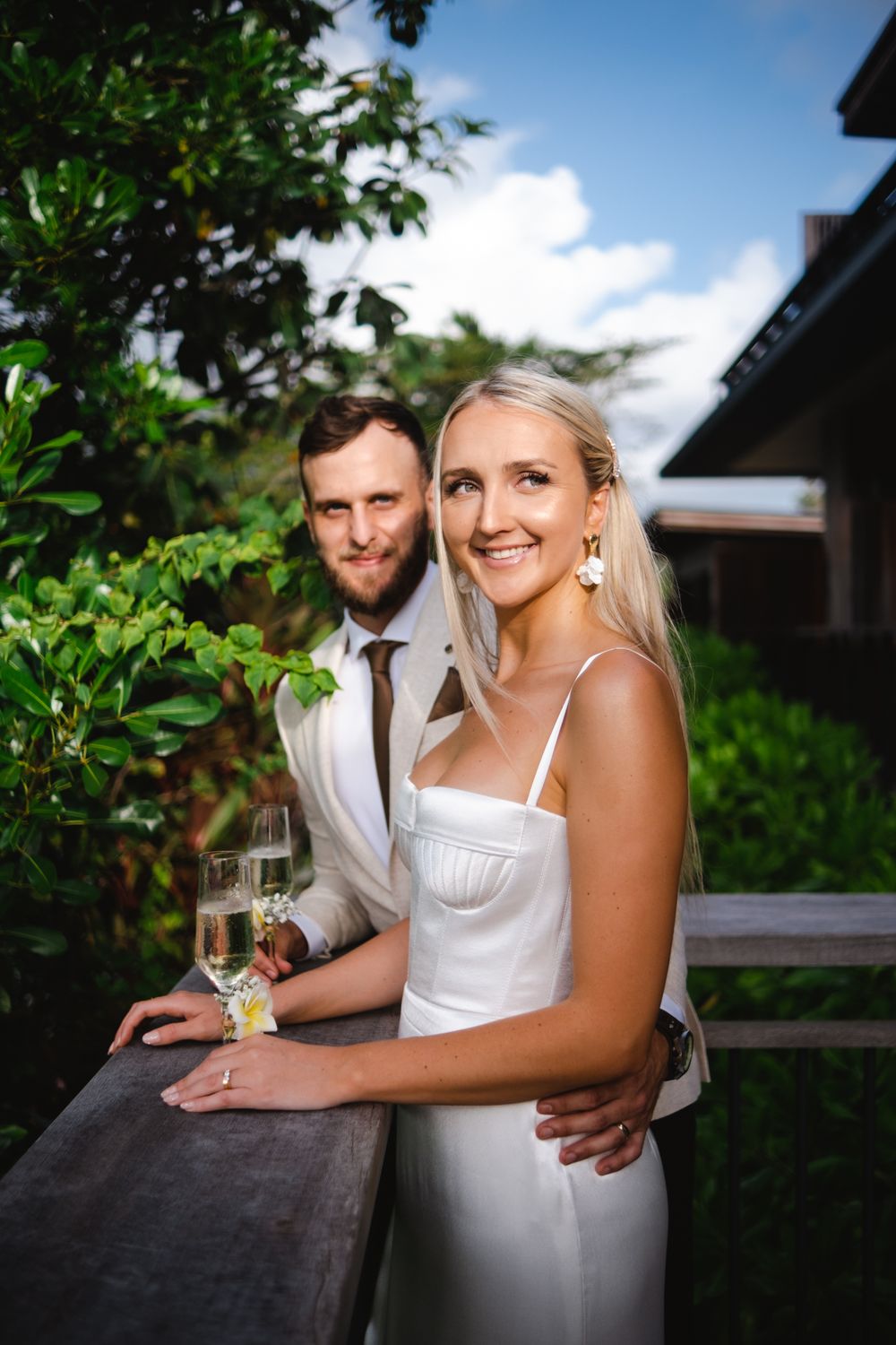 Smiling bride in white gown poses with groom holding champagne on garden terrace.