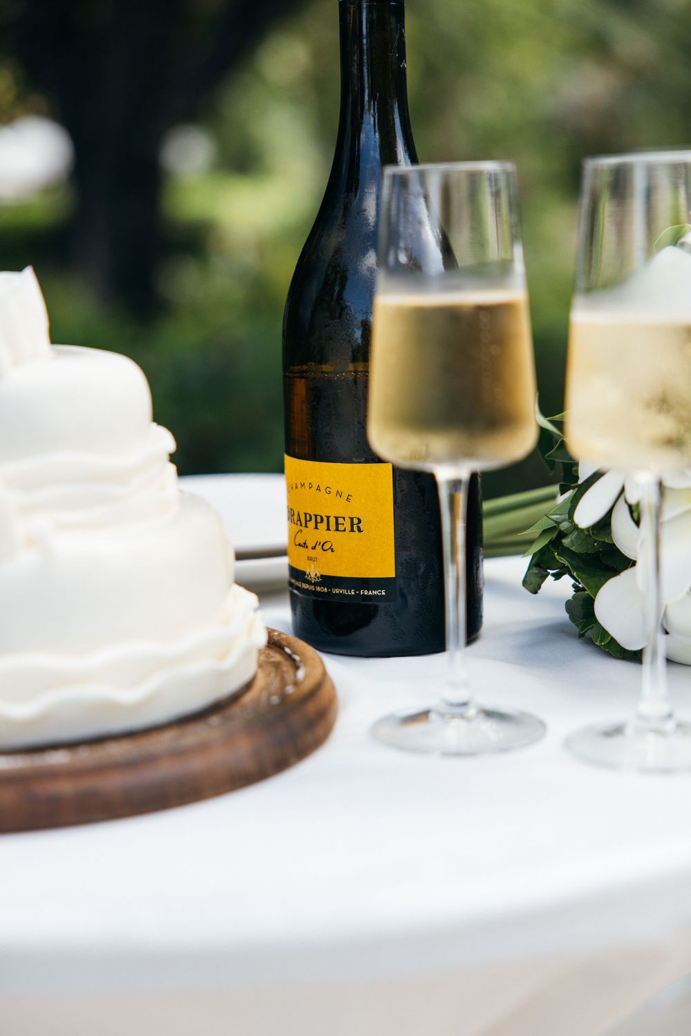 Wedding table with white tiered cake, Drappier champagne bottle and filled flutes outdoors.