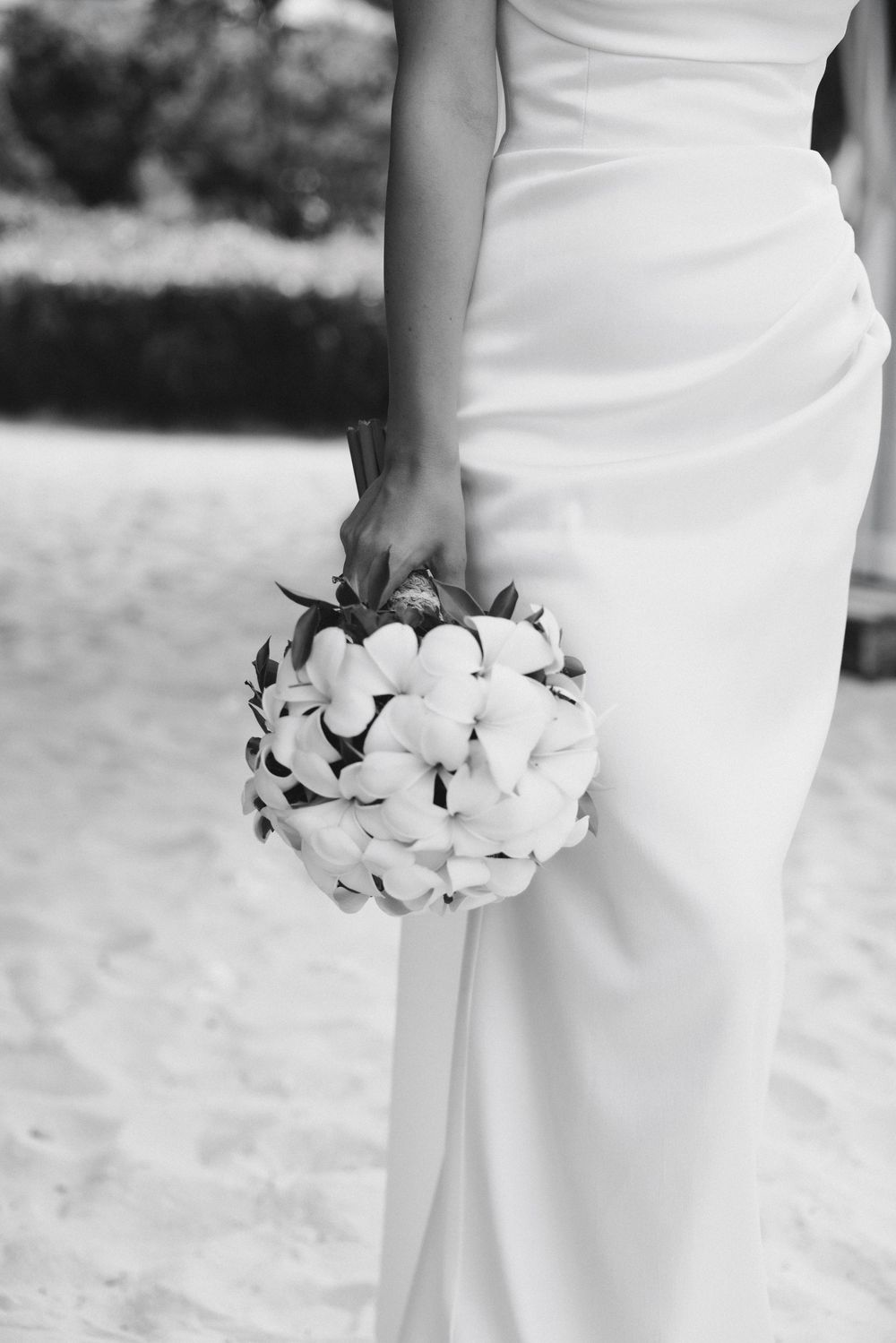 Bride in white gown holds round tropical floral bouquet on sandy beach in black and white.