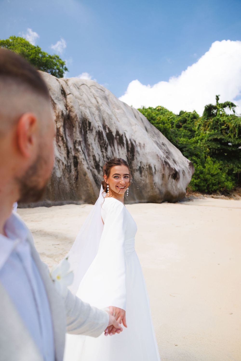 Smiling bride in white gown holds grooms hand on a sandy beach with large rocks in background.