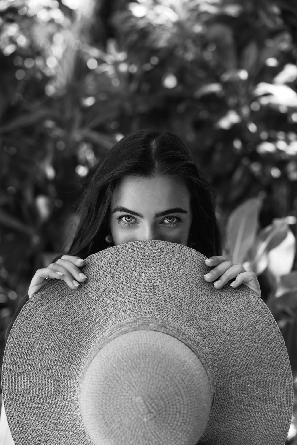 Beautiful woman with striking eyes peeks over a wide-brim straw hat surrounded by tropical foliage.