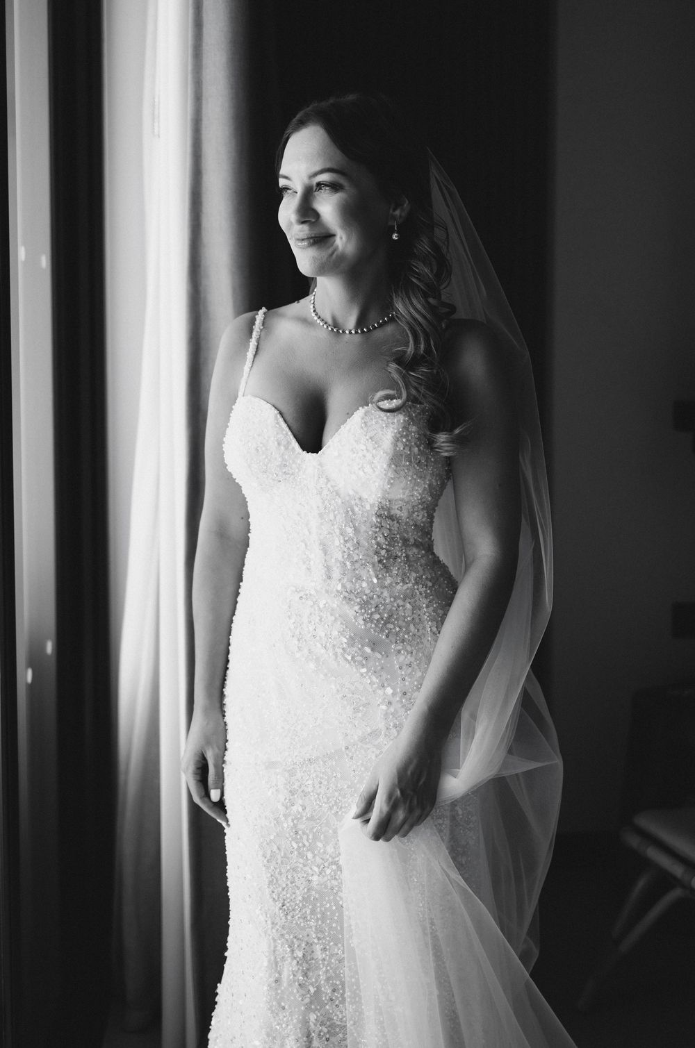 Elegant bride in beaded mermaid gown and veil smiles softly while standing by a window.