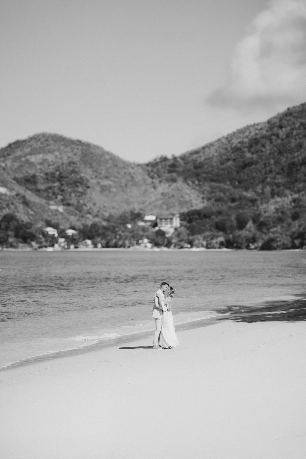 Bride and groom embrace on a white sand beach with lush green mountains and ocean in background.