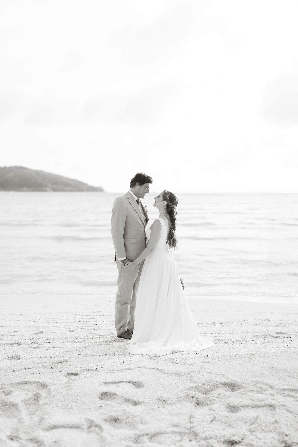 Bride and groom share a romantic moment on a sandy beach by the ocean in black and white photo.