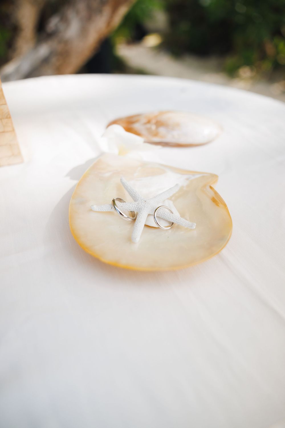 Wedding rings resting on a starfish inside a large seashell on a white table at a beach ceremony.