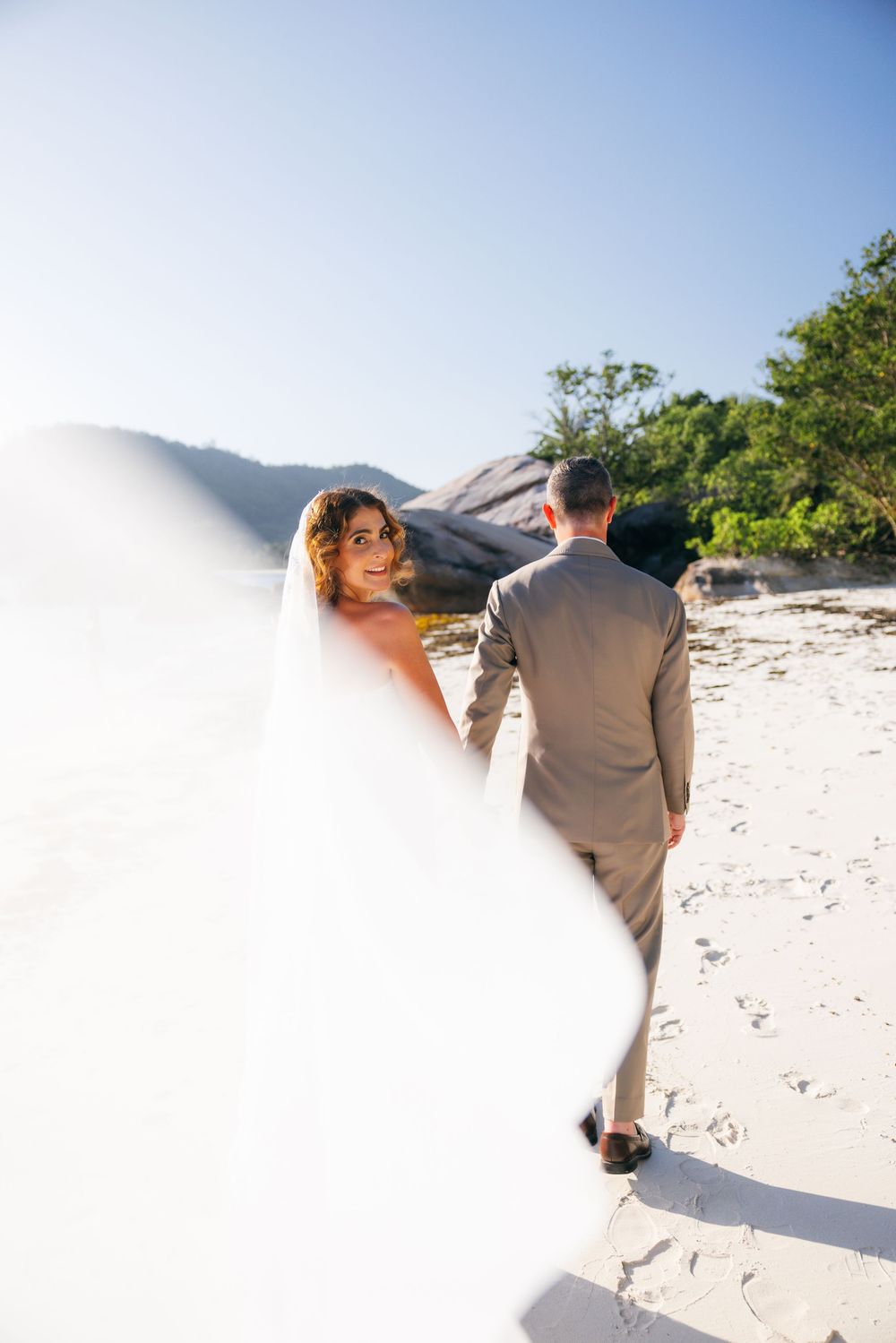 Bride glances back as her veil billows in the wind while walking with groom on a sunny beach.