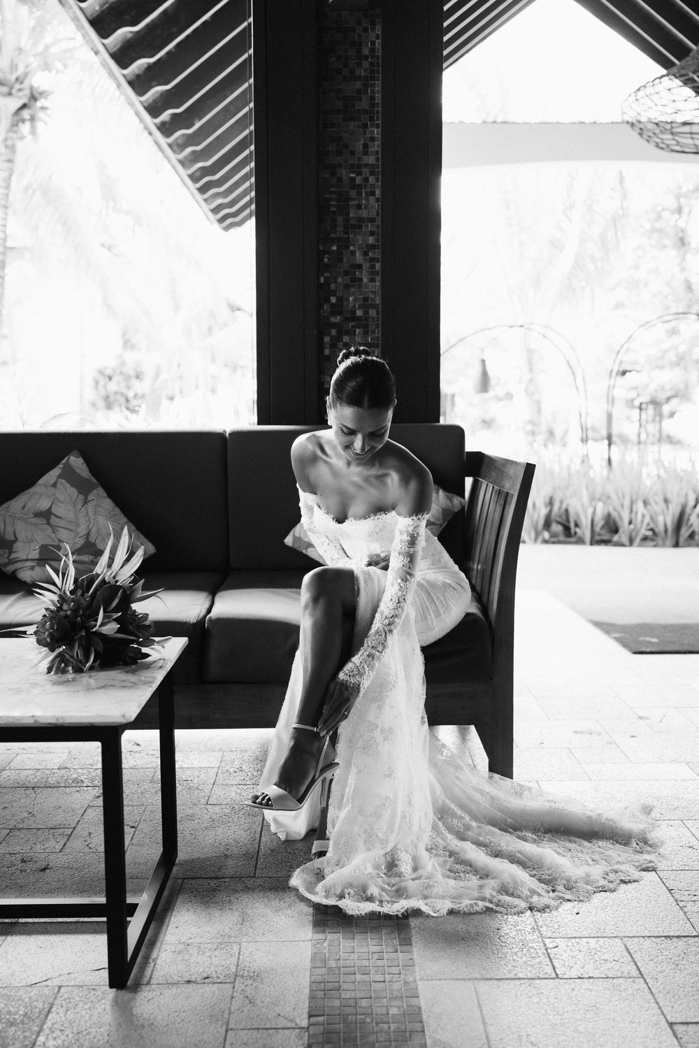 Bride in lace wedding gown fastens her heels while seated in a tropical villa in black and white.