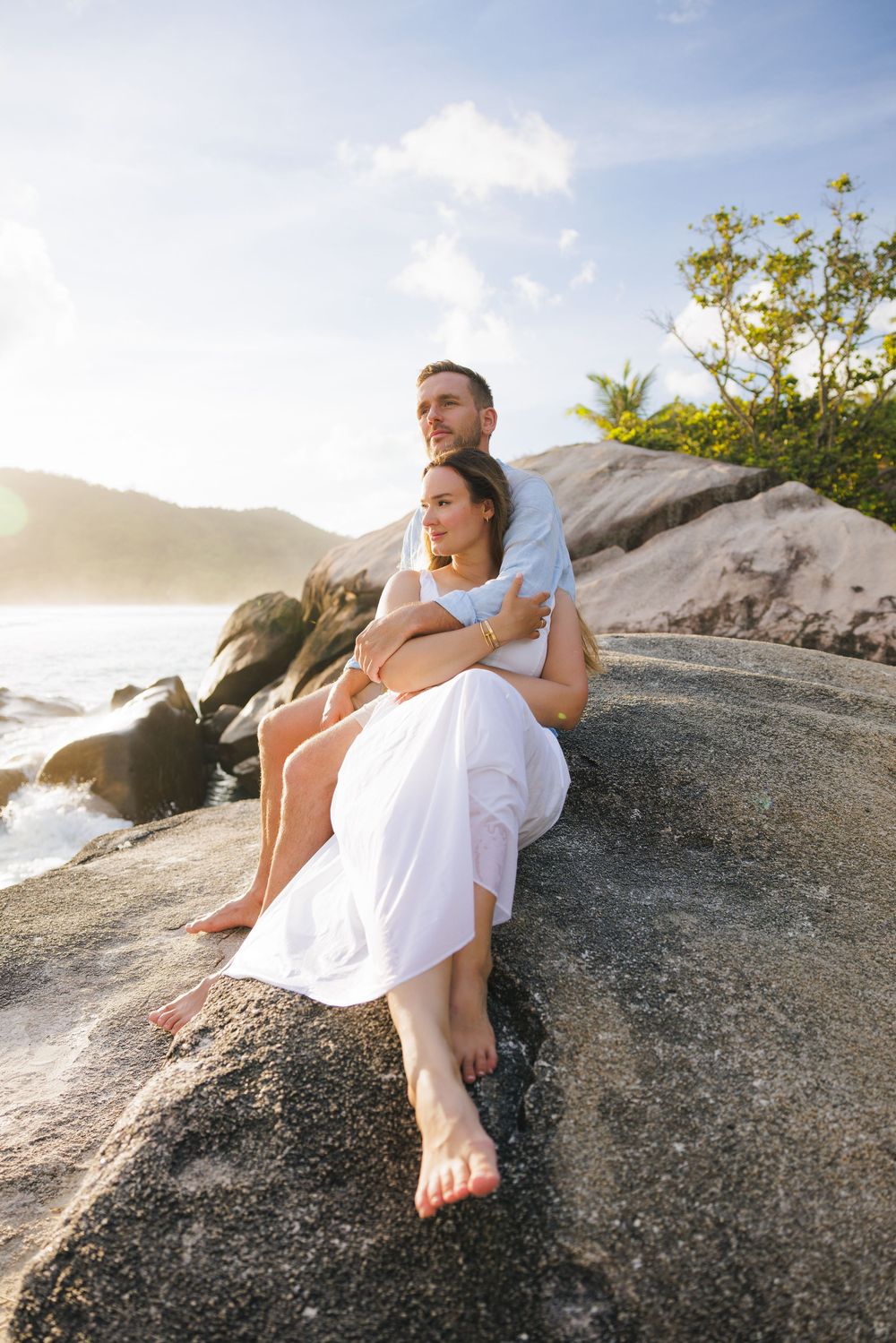 Couple embraces on coastal rocks at sunset, bride in white dress seated in grooms lap.