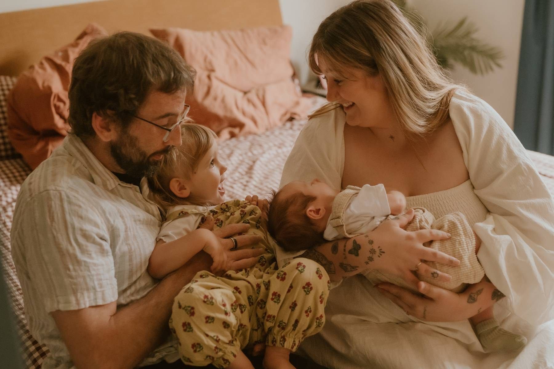 A happy family of four sits together on a bed, parents lovingly holding their toddler and newborn baby.