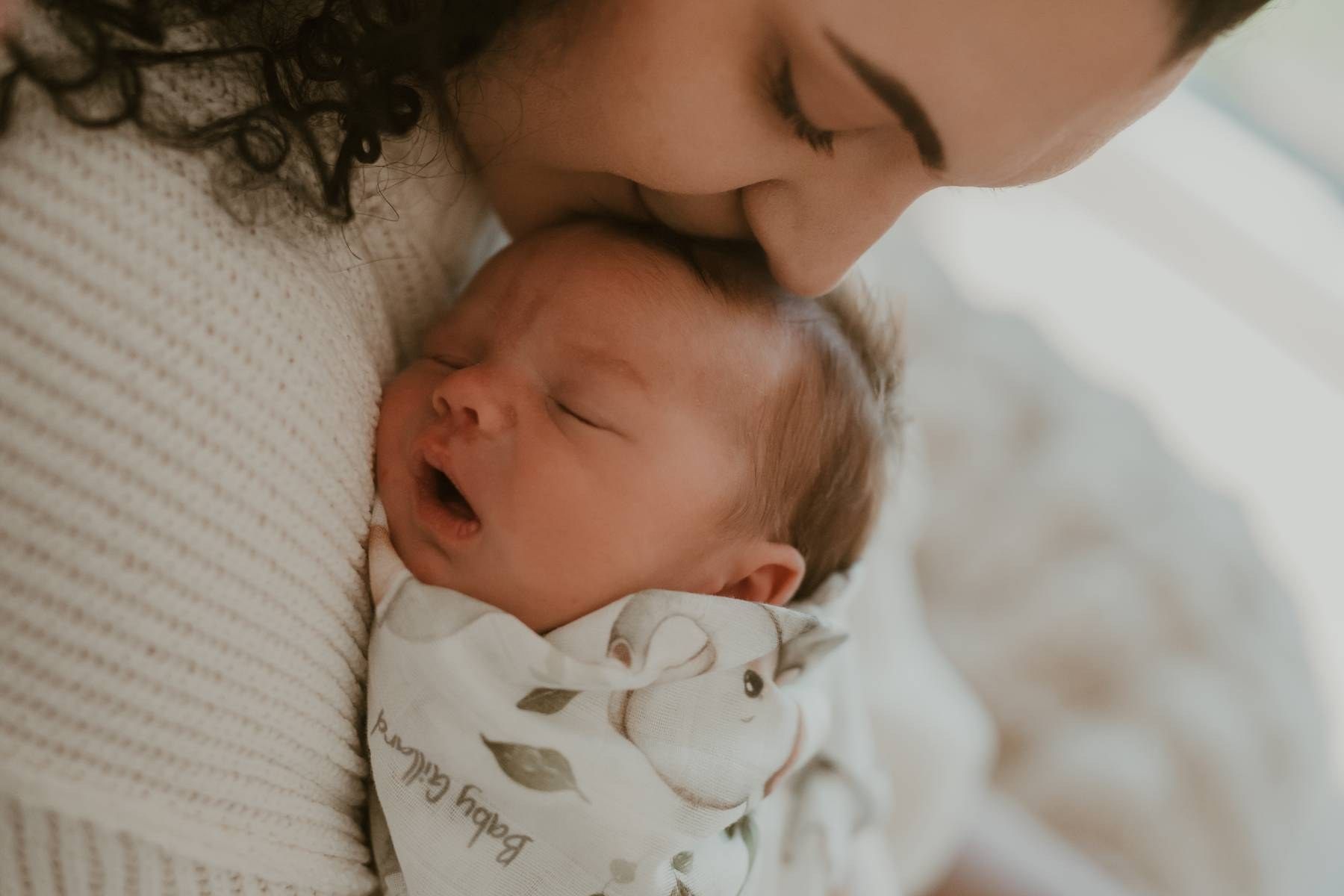 A mother tenderly kisses her sleeping newborn baby wrapped in a floral swaddle blanket.