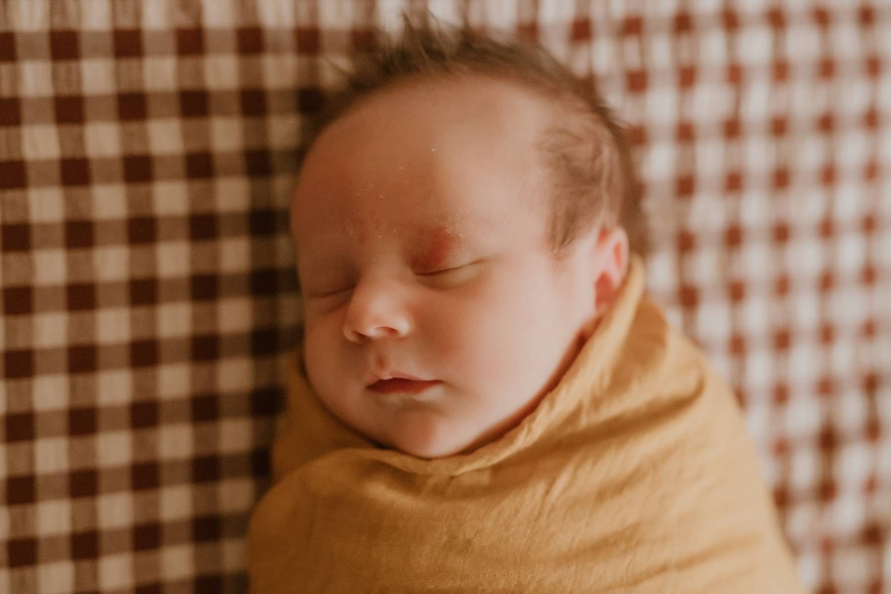 Peaceful newborn baby swaddled in a mustard yellow wrap, sleeping on a brown gingham blanket.