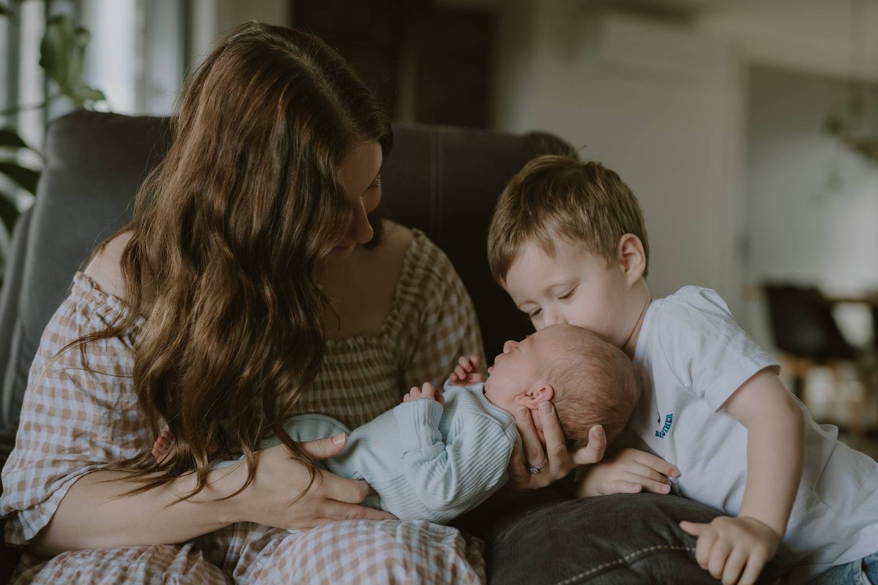 A mother holds her newborn baby while her young toddler son gently kisses the infant in a cozy living room.