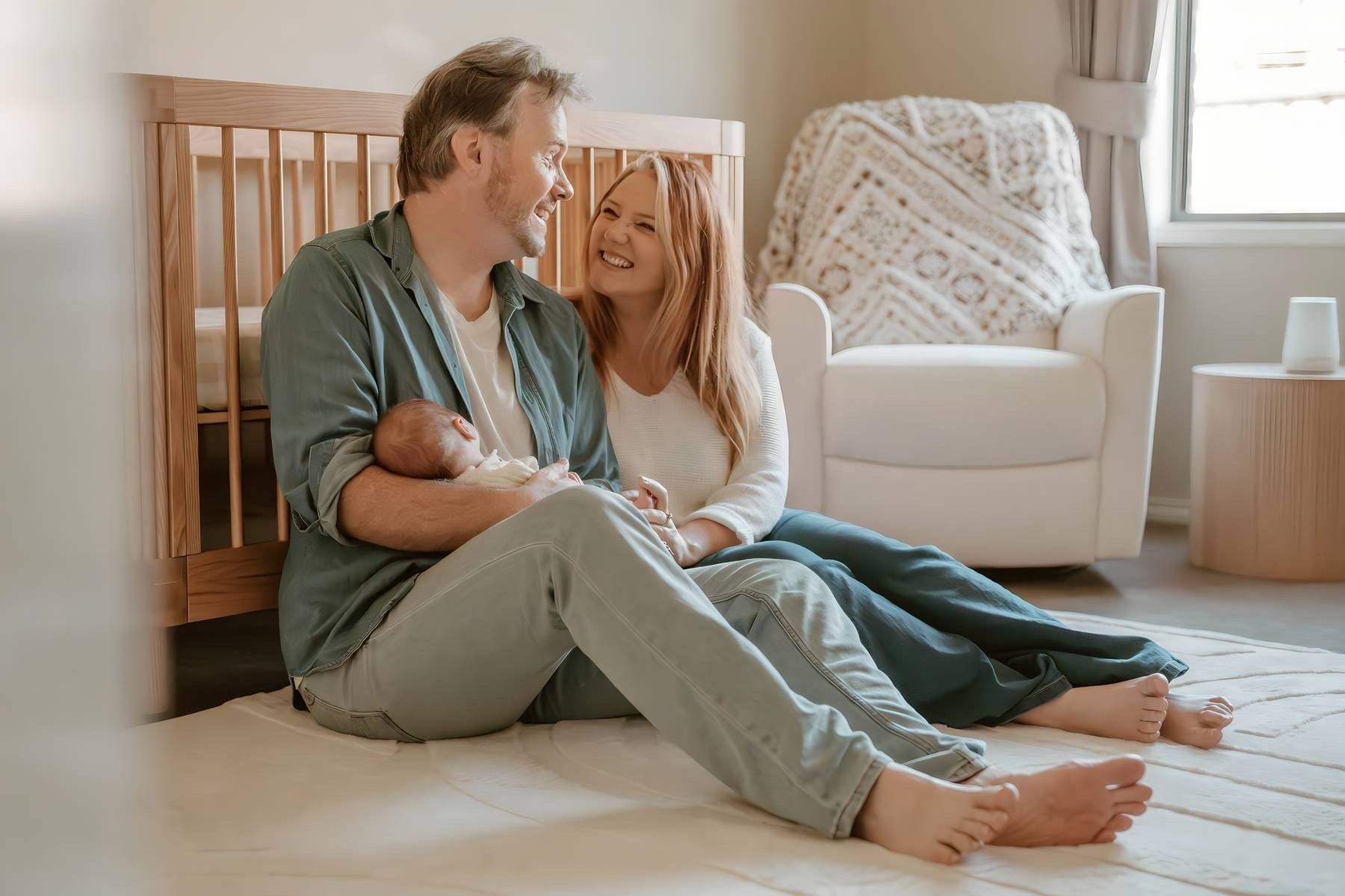 A couple sits on a nursery floor smiling, with the man holding a newborn baby near a crib and white rocking chair.