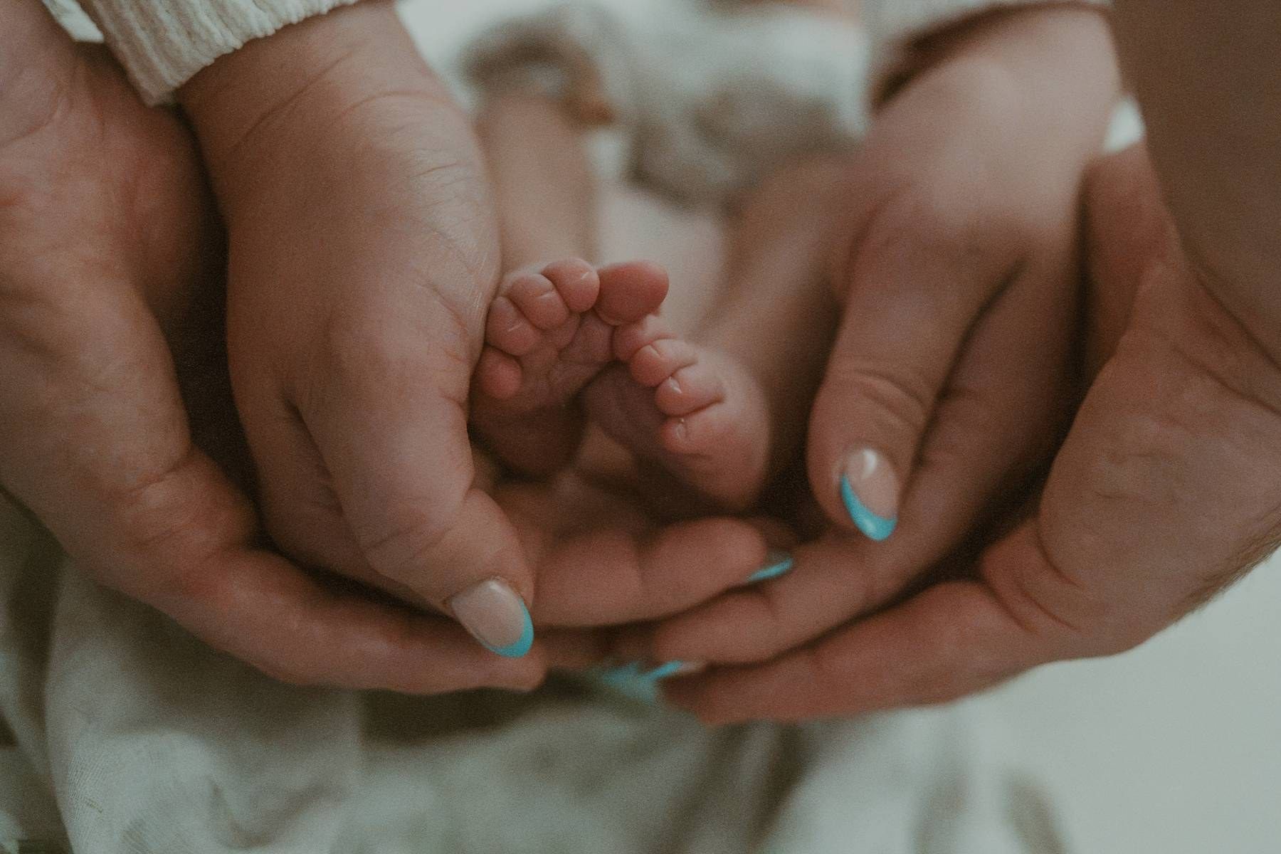 Adult hands cradling tiny newborn baby feet, with light blue painted nails visible, capturing a tender family moment.
