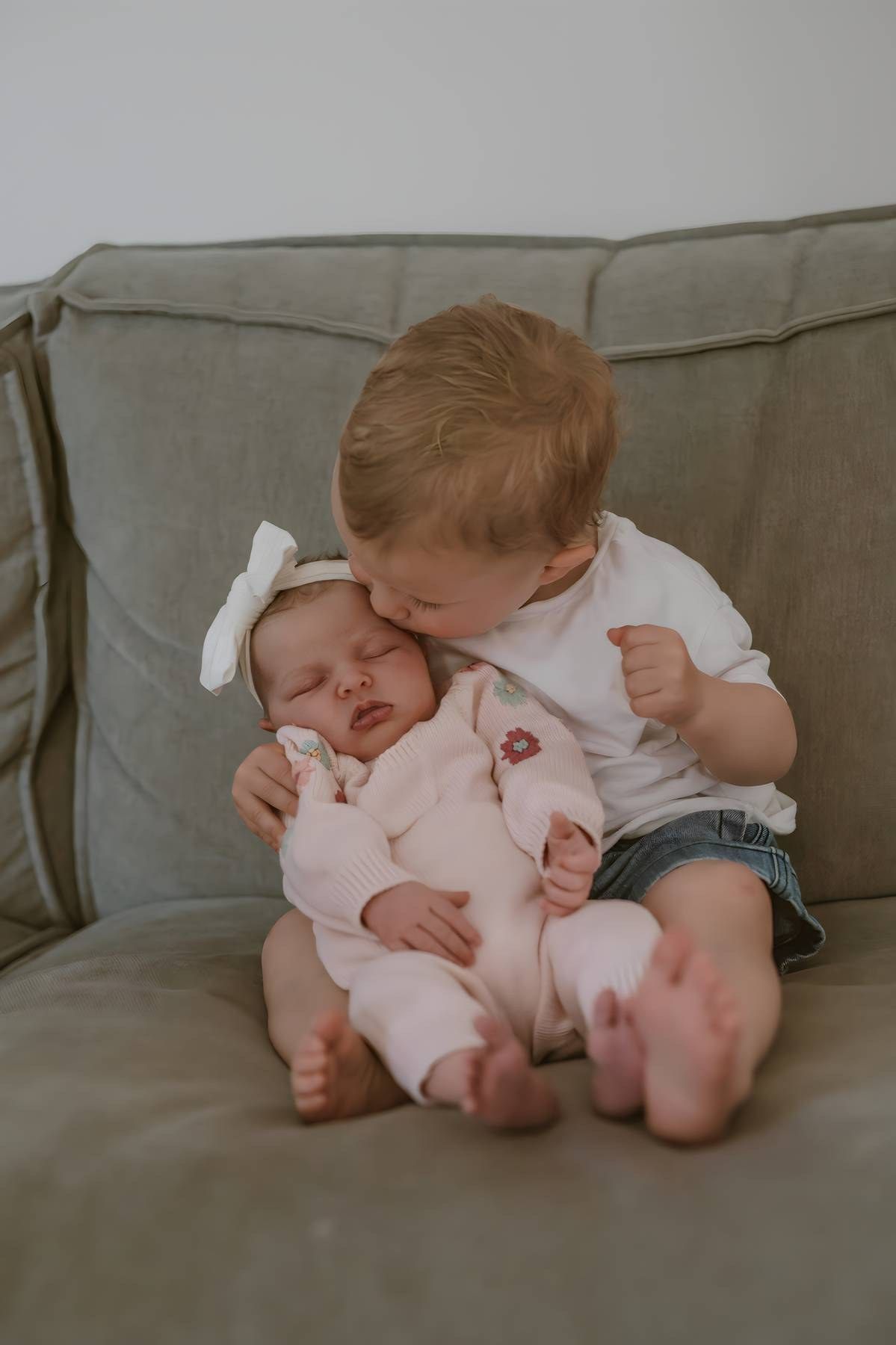 A toddler in a white shirt gently kisses a sleeping newborn baby in a pink outfit on a gray couch.
